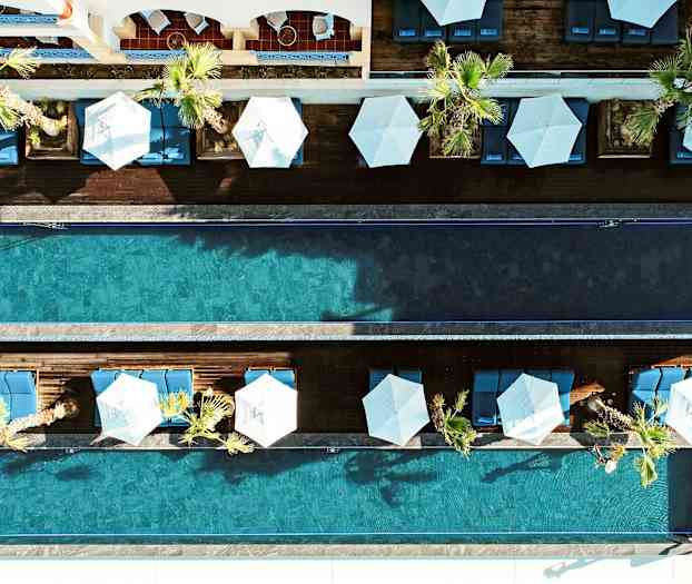 En fågelperspektivbild av två parallella simbassänger omgivna av palmer, solstolar och parasoller. (AI-genererad text)