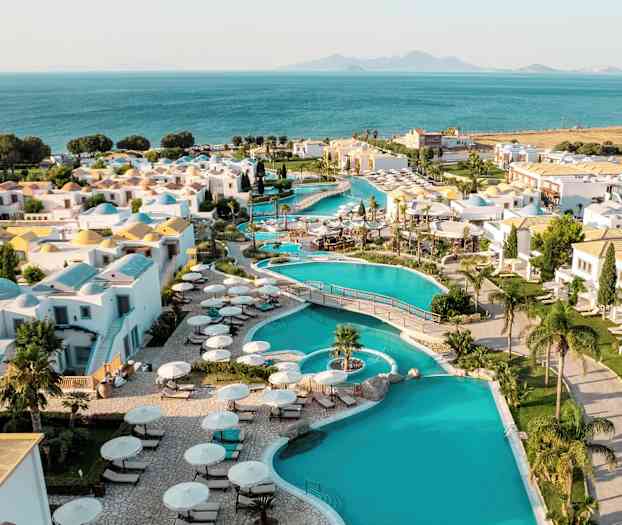 Vy över ett semesterkomplex med pooler, vita byggnader och utsikt mot havet i bakgrunden. (AI-genererad text)