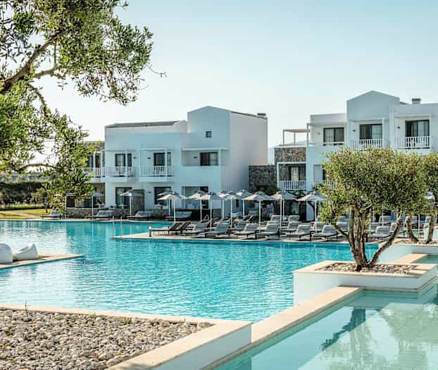 Bilden visar ett hotell med vit fasad vid en stor pool, omgiven av solstolar och parasoller samt grönska. (AI-genererad text)