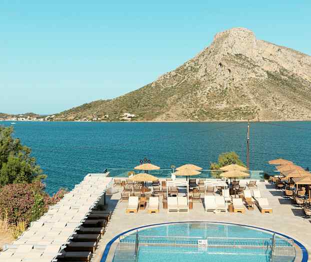 Utsikt över en pool, en terrass med solstolar, och en havsmiljö med berg i bakgrunden. (AI-genererad text)