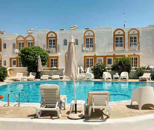 En hotellpool med solstolar och parasoll framför en byggnad med vita fasader och orange detaljer. (AI-genererad text)