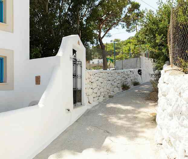 Vit byggnad med blå fönsterramar och en järngrind vid en stenmur. Solig gångväg omgiven av träd. (AI-genererad text)