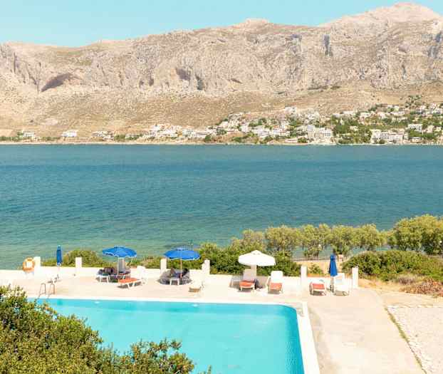 Utsikt över en pool med solstolar och parasoller, med havet och berg i bakgrunden. (AI-genererad text)