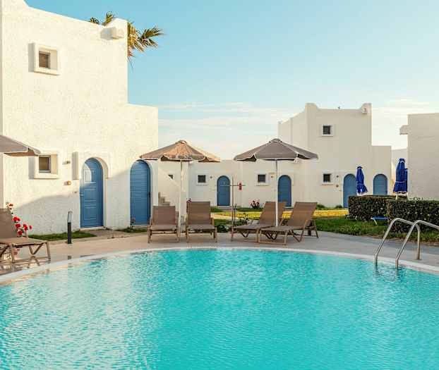 En pool omgiven av solstolar och parasoller, med vita byggnader med blå dörrar i bakgrunden. (AI-genererad text)