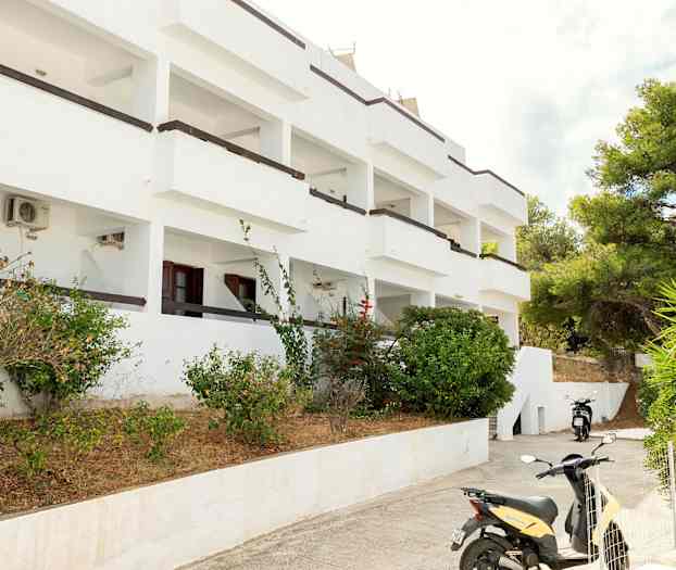 Vit byggnad med balkonger omgiven av buskar och en parkerad motorcykel. (AI-genererad text)