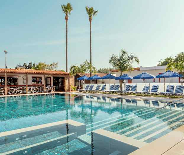 En pool omgiven av solstolar och parasoller med en bar i bakgrunden. (AI-genererad text)