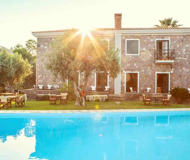 En stenbyggnad bakom en pool med trädgårdsmöbler och träd. (AI-genererad text)