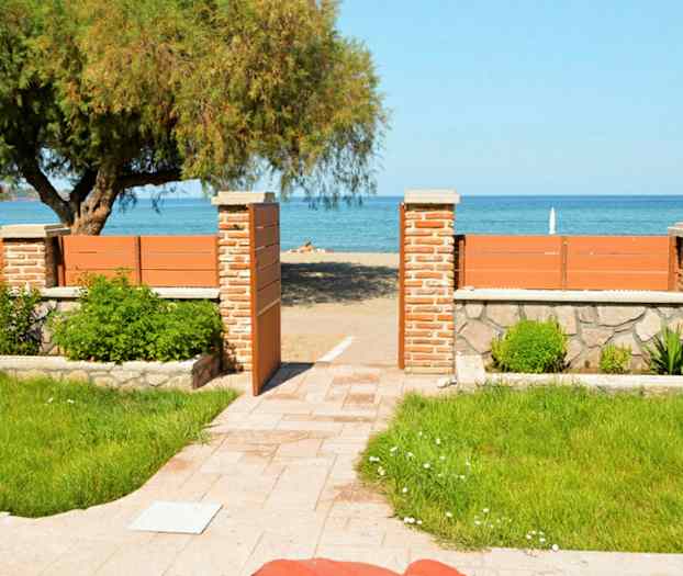 En grön trädgård med stenbelagd gång leder till en strand bakom ett trästaket med en öppnad grind. (AI-genererad text)