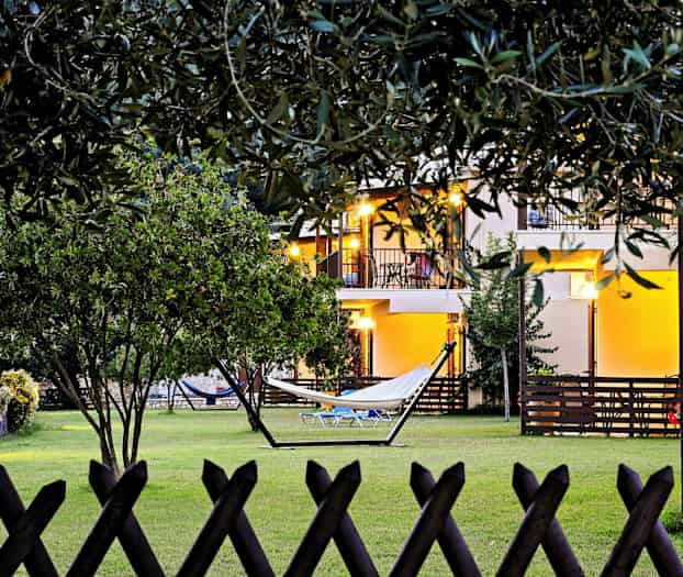 Bild av en trädgård med gräsmatta, hängmatta och omgivande vegetation. I bakgrunden syns ett hus med upplysta balkonger. (AI-genererad text)