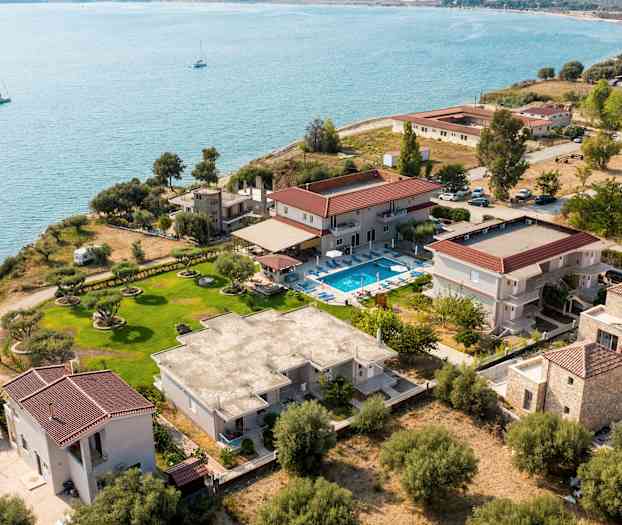 Fågelperspektiv över en kustfastighet med olika byggnader, en gräsmatta och en pool nära vattnet. (AI-genererad text)