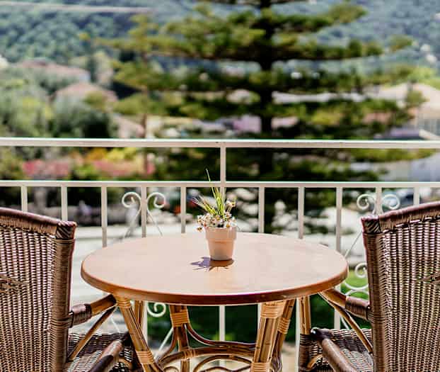 En balkong med ett träbord och två rottingstolar, och en liten blomkruka med blommor på bordet. I bakgrunden syns träd och byggnader. (AI-genererad text)