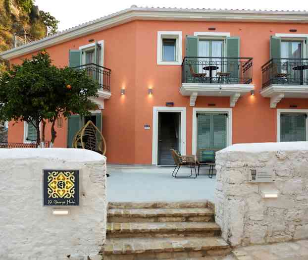 En liten hotellbyggnad med orange fasad och gröna fönsterluckor samt balkonger. Framför byggnaden finns en sittgrupp och ett träd med frukt. (AI-genererad text)