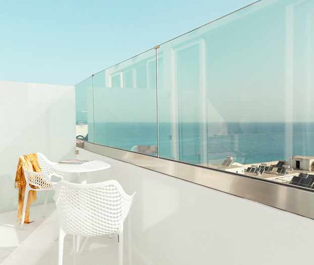 En vit balkong med två stolar och ett bord, med utsikt över havet och byggnader. (AI-genererad text)