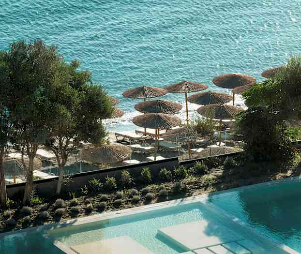 Vy över poolen, träd och stranden med parasoller och solsängar mot havet. (AI-genererad text)