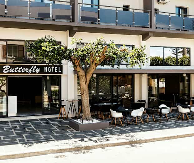 Framsidan av ett hotell med skylt texten "Butterfly Hotel", en uteplats med bord och stolar samt ett träd står framför byggnaden. (AI-genererad text)