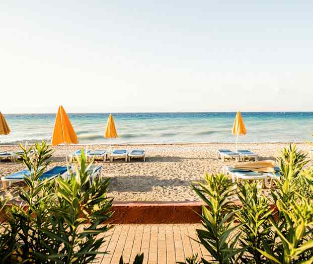 En strand med solstolar och gula parasoller vid ett blått hav, omgiven av växter. (AI-genererad text)