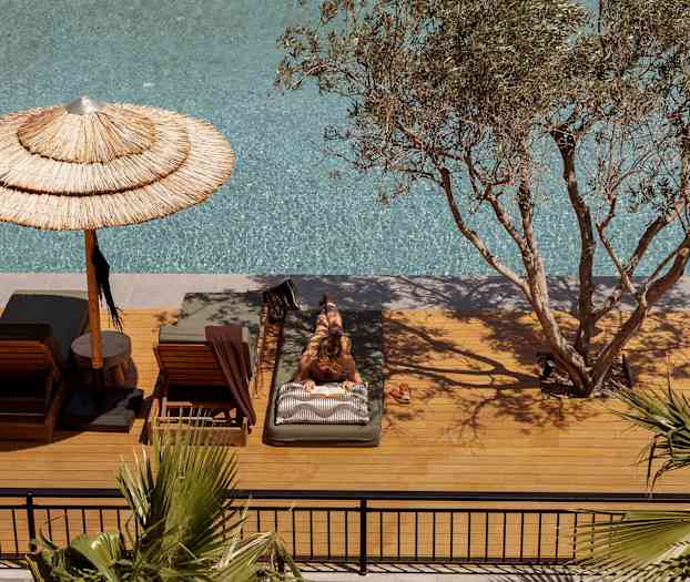 Person som ligger på en solbädd vid en pool, omgiven av träd och parasoll. (AI-genererad text)