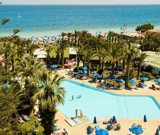 En pool omgiven av solstolar och palmer, med en sandstrand och havet i bakgrunden. (AI-genererad text)