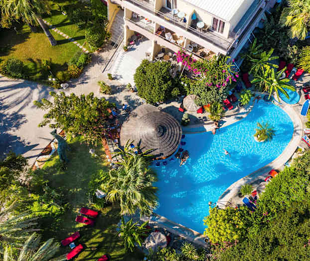 Vy ovanifrån av en poolområde omgiven av grönska och ett hotell. (AI-genererad text)