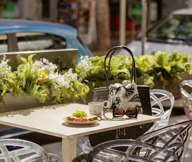 Bord med croissant, kopp och solglasögon på uteservering med blomsterdekorationer. (AI-genererad text)