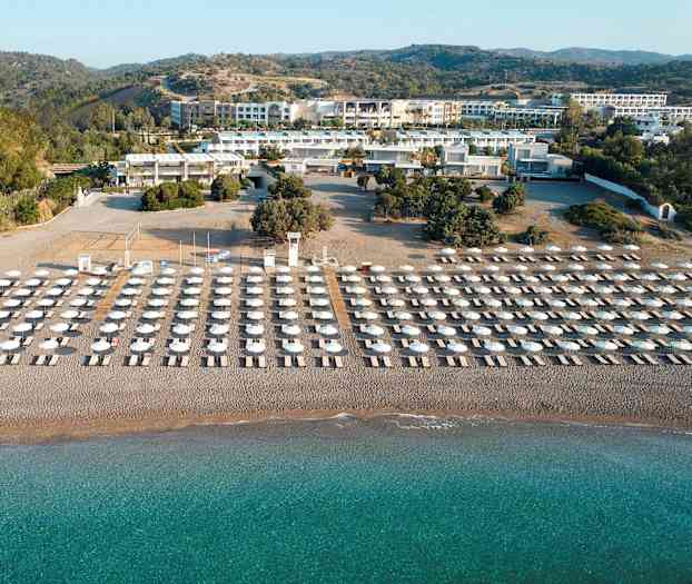 Solstolar och parasoller i flera rader på en sandstrand med turkost vatten, omgivna av grönska och byggnader i bakgrunden. (AI-genererad text)