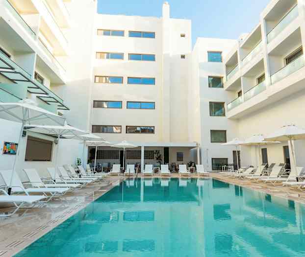 Bild av ett hotellområde med en pool omgiven av solstolar och parasoller ovanför. Bakgrunden visar vita byggnader med balkonger. (AI-genererad text)