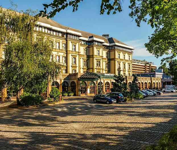 En bild av en byggnad med texten 'Grand Hotel' på fasaden, omgiven av träd och parkerade bilar. (AI-genererad text)