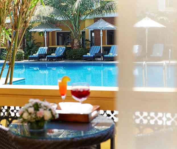 Bilden visar en pool med solstolar och parasoller samt en närbild på ett bord med drycker och blommor i förgrunden. (AI-genererad text)