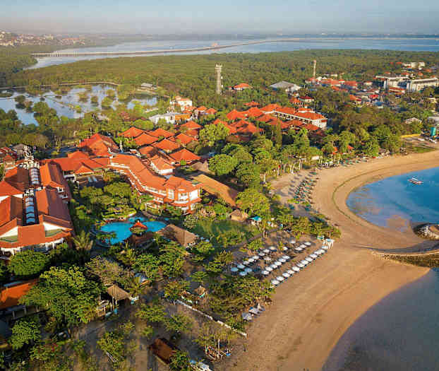 Luftbild över ett strandhotell med omgivande tropisk vegetationen, stranden med solsängar och havet. (AI-genererad text)