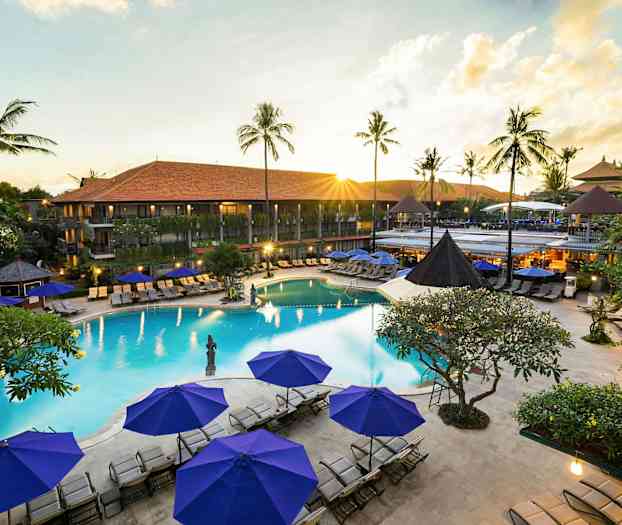 Bild av en resort med en pool, omgivande solstolar och parasoll samt gröna palmer i bakgrunden. (AI-genererad text)