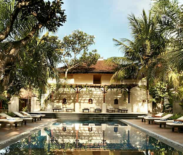 En utomhuspool omgiven av solstolar och träd med en byggnad i bakgrunden. (AI-genererad text)