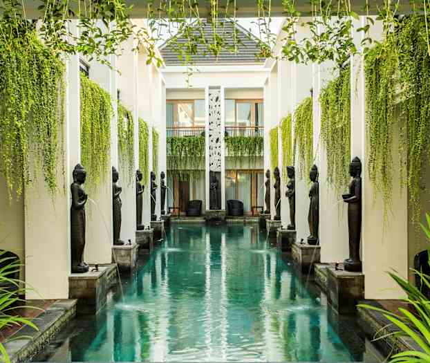 En lång, smal pool omgiven av pelare, statyer och överhängande grönska. (AI-genererad text)