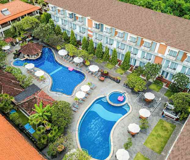 En fågelperspektiv över ett hotellområde med två pooler, solstolar och parasoller, omgivna av växtlighet och byggnader. (AI-genererad text)