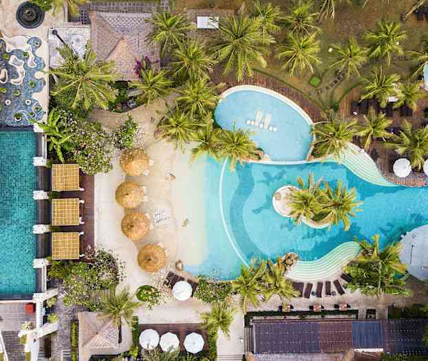 Flygfoto av en poolanläggning med solstolar, parasoller och frodiga palmsällskap. (AI-genererad text)