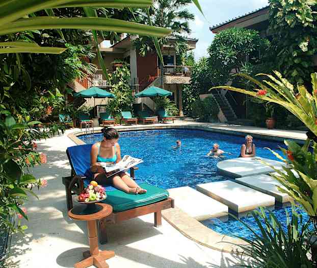 Ett poolområde med solstolar, parasoller och grönskande växter. En person sitter i en solstol intill bord med frukter, medan andra badar i poolen. (AI-genererad text)