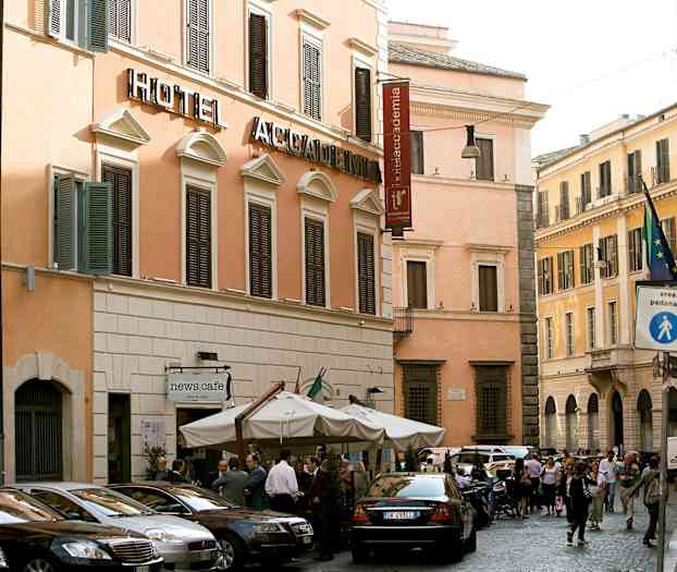 En gatuscen med en hotellbyggnad med beige fasad och mörka fönsterluckor. Framför byggnaden står flera parkerade bilar och ett kafé med parasoller. (AI-genererad text)