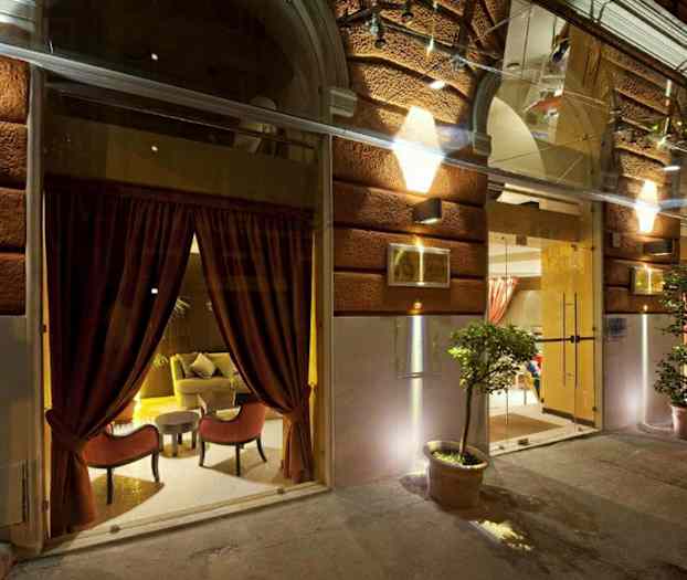 Bild på ingången till en byggnad med en upplyst fasad, stora glasfönster med röda gardiner och krukväxter. (AI-genererad text)