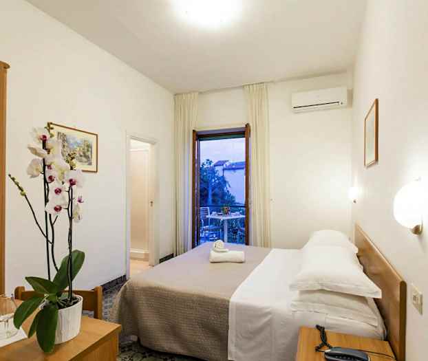 Ett hotellrum med en dubbelsäng, nattduksbord, en blomvas på ett litet bord och en balkong med utsikt. (AI-genererad text)
