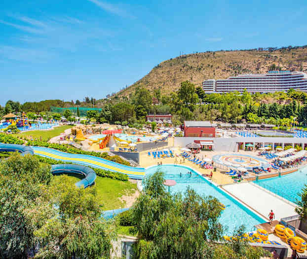 Bilden visar ett stort utomhusvattenland med pooler, vattenrutschkanor och omgivande solstolar. En stor byggnad och sluttande terräng syns i bakgrunden. (AI-genererad text)