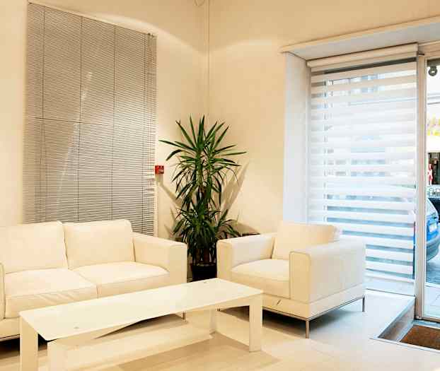 Ett ljust vardagsrum med vita möbler, en växt och stora fönster med persienner. (AI-genererad text)
