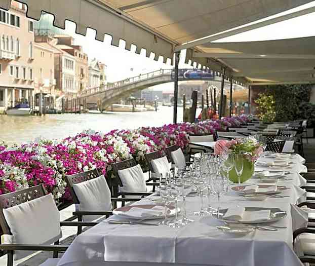 Ett uppdukat matbord med vita dukar placerat under ett soltak vid en kanal fylld med båtar och dekorerad med färgglada blommor längs kanten. (AI-genererad text)