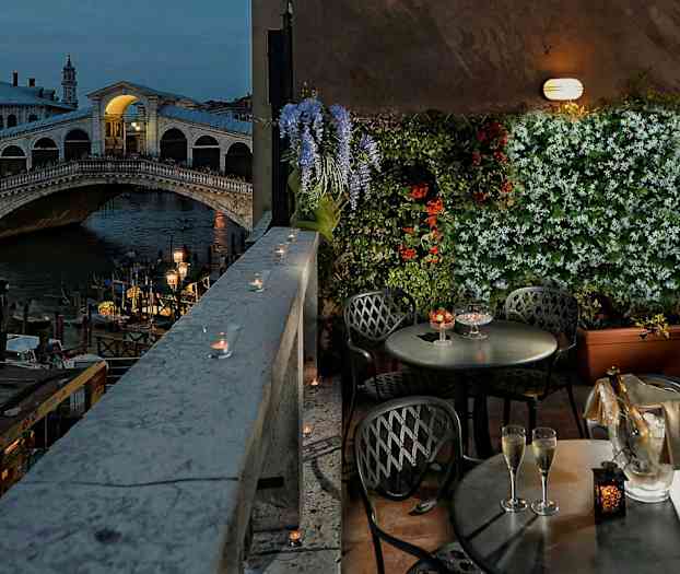 Bilden visar en terrass med bord och stolar, utsikt över Rialtobron i Venedig, blommor och ljus dekorerar området. (AI-genererad text)