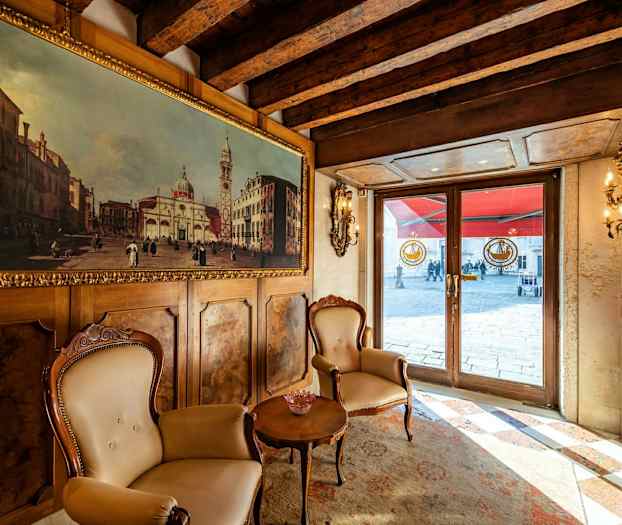 Inomhusscen med två stolar och ett bord framför en vägg med en målning, med utsikt mot en exteriör genom glasdörrar. (AI-genererad text)