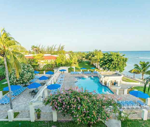 Bild av ett hotellområde vid havet med pool, solstolar och parasoller omgivna av grönska och palmer. (AI-genererad text)