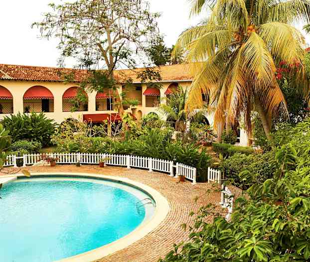 En rund pool omgiven av en stenbelagd yta och vita staket. I bakgrunden finns en byggnad med valvmålade fönster och röda markiser samt gröna omgivande växter. (AI-genererad text)