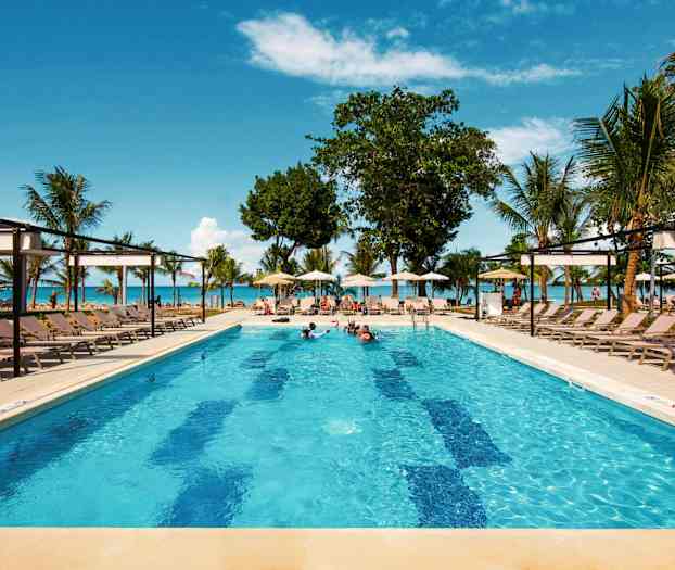 En pool med solstolar omkring, omgiven av palmer och med en utsikt mot havet och blå himmel. (AI-genererad text)