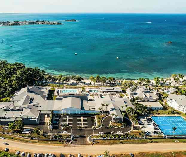 Flygfoto av en kustresort med pooler, byggnader och en tennisbana vid havet. (AI-genererad text)
