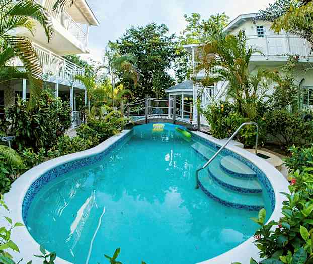 En utomhuspool omgiven av gröna växter med byggnader i bakgrunden. (AI-genererad text)