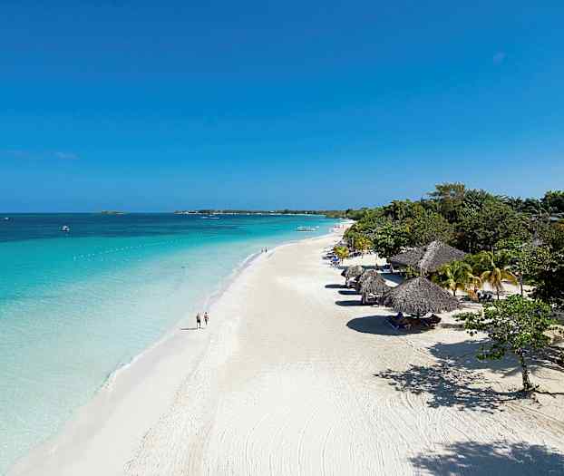 En vit sandstrand med parasolltak och klart blått vatten, omgiven av grönska. (AI-genererad text)