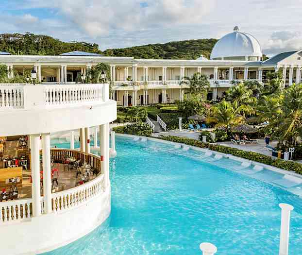 En stor byggnad med vita pelare och kupol vid en pool omgiven av palmer och solstolar. (AI-genererad text)